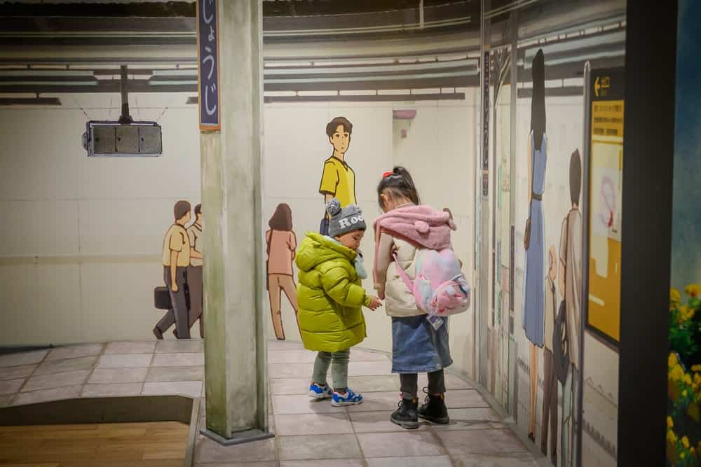 日本愛知-吉卜力公園｜兩個小女孩，綠色外套，淺藍色裙子，室內，牆壁，柱子，地板，動漫人物，廣告牌。