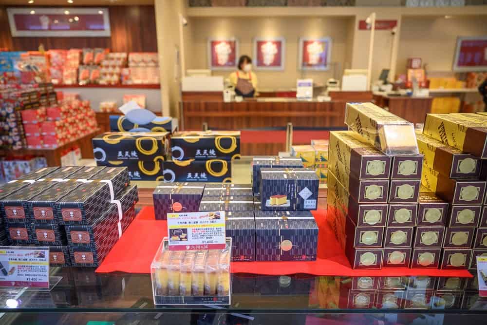 日本長崎-豪斯登堡｜店內，玻璃櫃檯，包裝食品，紅色檯面，價目表，收銀台，陳列架，顧客。