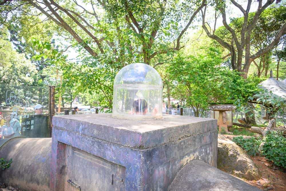 桃園六福村墓碑鎮｜石台、透明塑膠罩、樹、石燈籠、石塊、欄杆、圍牆。