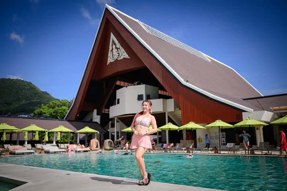 泰國普吉-Clubmed｜一位身穿比基尼的女性，站在泳池邊，背景是具有獨特屋頂的建築物，周圍有綠色陽傘和躺椅，泳池中有其他人在