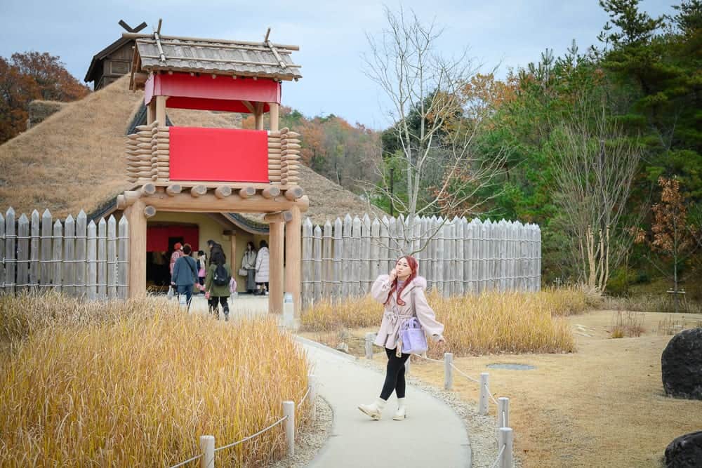 日本愛知-吉卜力公園｜建築物、登入口、木造圍欄、稻草屋頂、紅色牌匾、木制欄杆、步道、圍欄、樹木、黃色枯草、遊客、女子、草地