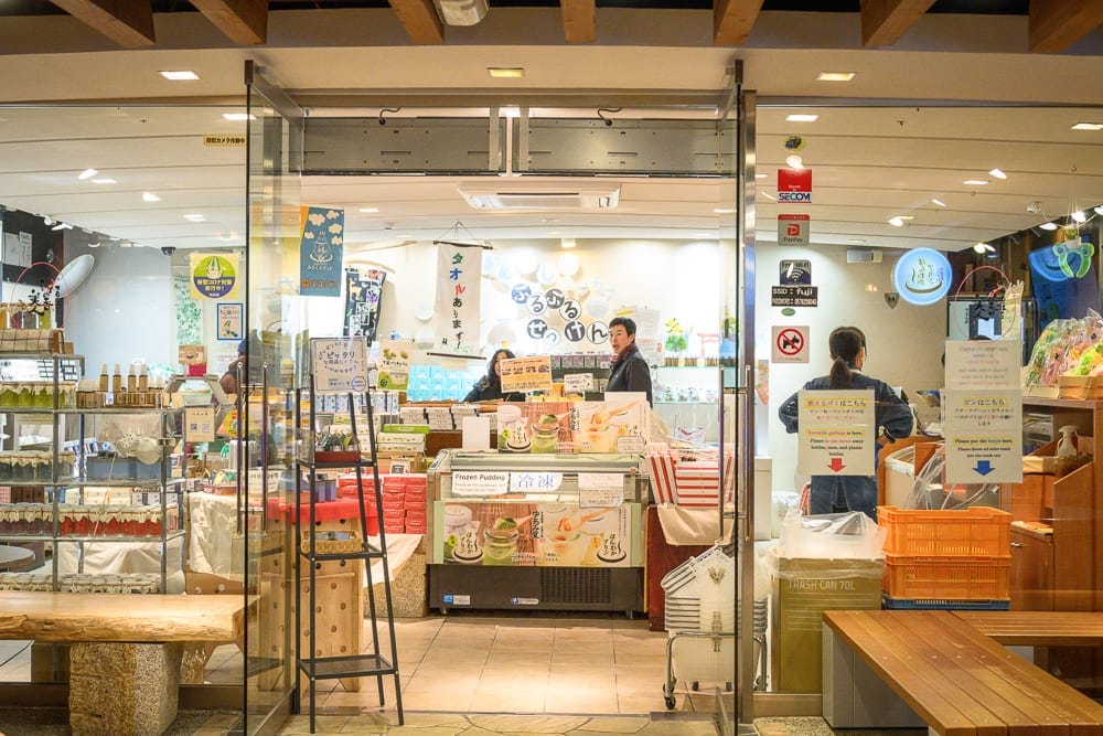 日本下呂-下呂老街散冊｜店內、玻璃門、貨架、食品、木桌，店員、顧客。室內燈光充足，陳列著各種食品和商品，貨架和玻璃櫃展示商品