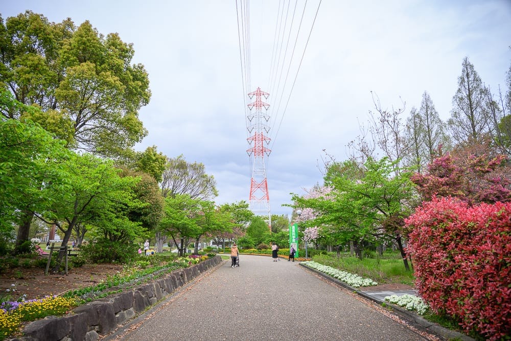 日本名古屋-戶田川綠地公園｜道路、樹、電塔、花壇、石牆、草坪、行人。