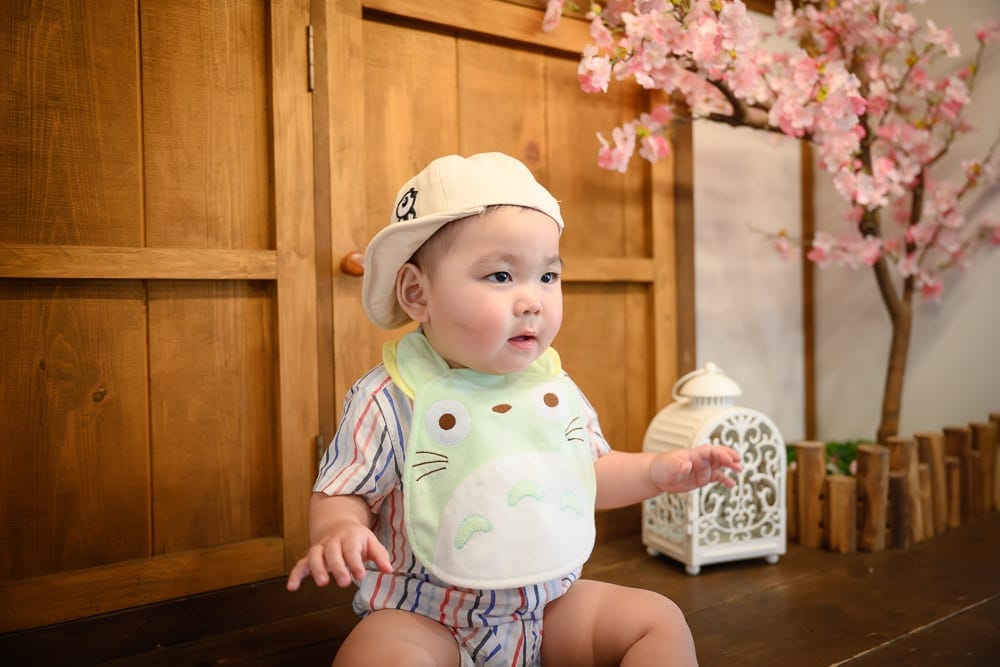 嘉義檜意森活村龍貓｜嬰兒、室內、木門、櫻花樹、燈籠、圍兜。
嬰兒穿著帽子和圍兜坐在木門前，旁邊有櫻花樹和燈籠。