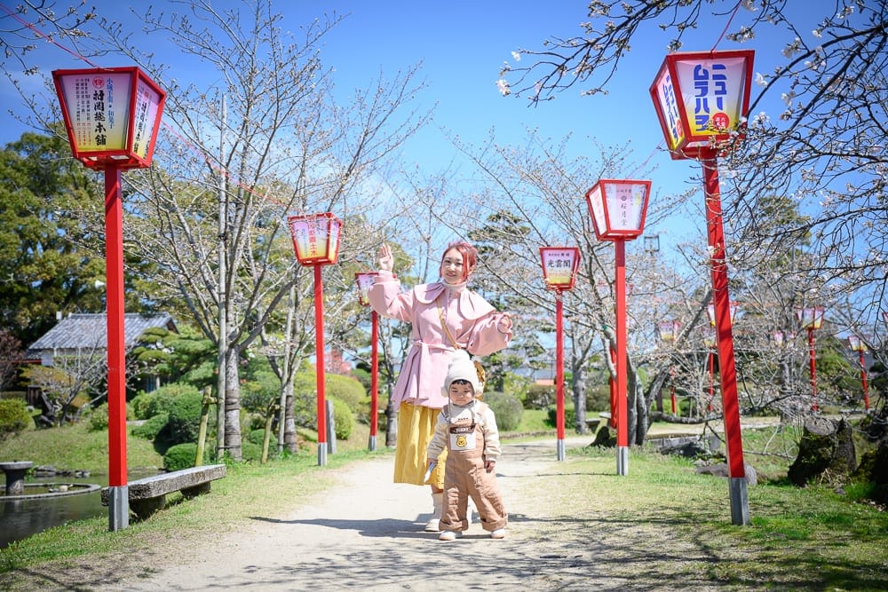 日本佐賀-小城公園櫻花｜紅色燈籠、樹、草地、建築物、道路、婦女、小孩。