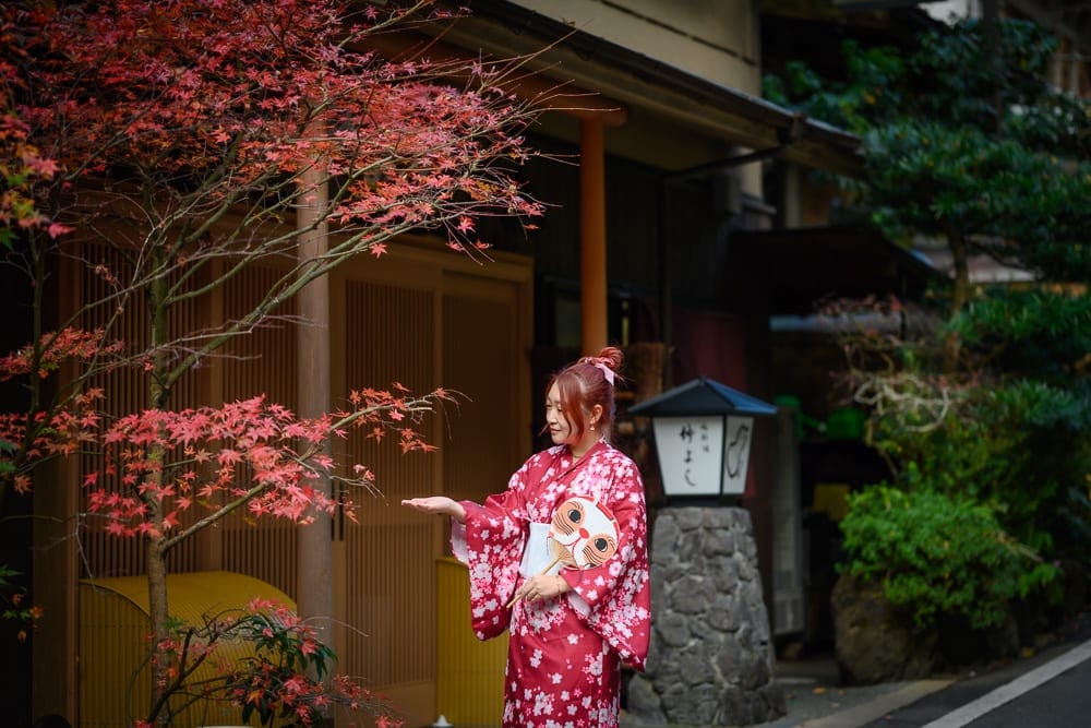 日本京都貴船神社｜紅葉樹、和服女子、傳統日式建築、石燈籠、綠植。
和服女子站立在紅葉樹前，伸手觸碰樹葉，背景是傳統的