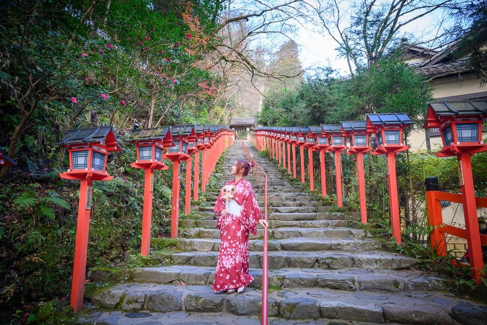 日本京都貴船神社｜石階，紅色燈籠，樹叢，女子和服。
一女子穿著紅色和服，挽著髮髻，提著拐杖，沿著佈滿青苔的石階而上，