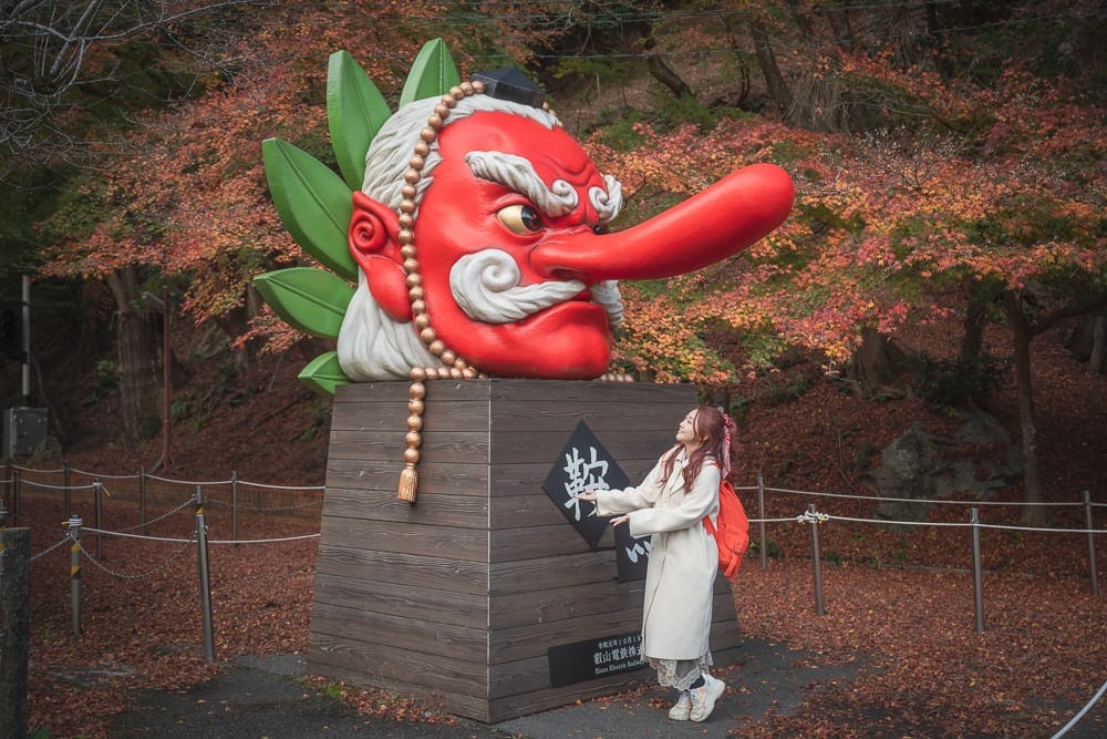 日本京都鞍馬寺修行之路｜巨大紅鼻子雕像、木製底座、女性、繩索圍欄、秋葉樹林。