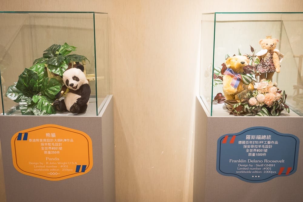 桃園小熊博物館｜兩個玻璃展示盒，熊貓玩偶、綠植，黃色標籤；熊、兔玩偶，花卉，藍色標籤。室內。