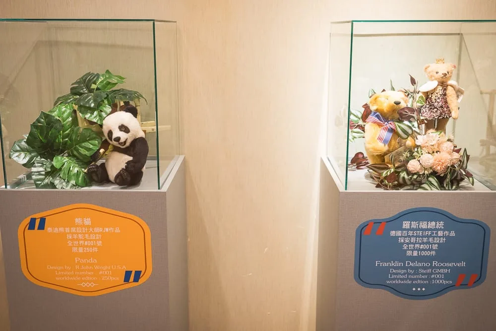 桃園小熊博物館｜兩個玻璃展示盒，熊貓玩偶、綠植，黃色標籤；熊、兔玩偶，花卉，藍色標籤。室內。