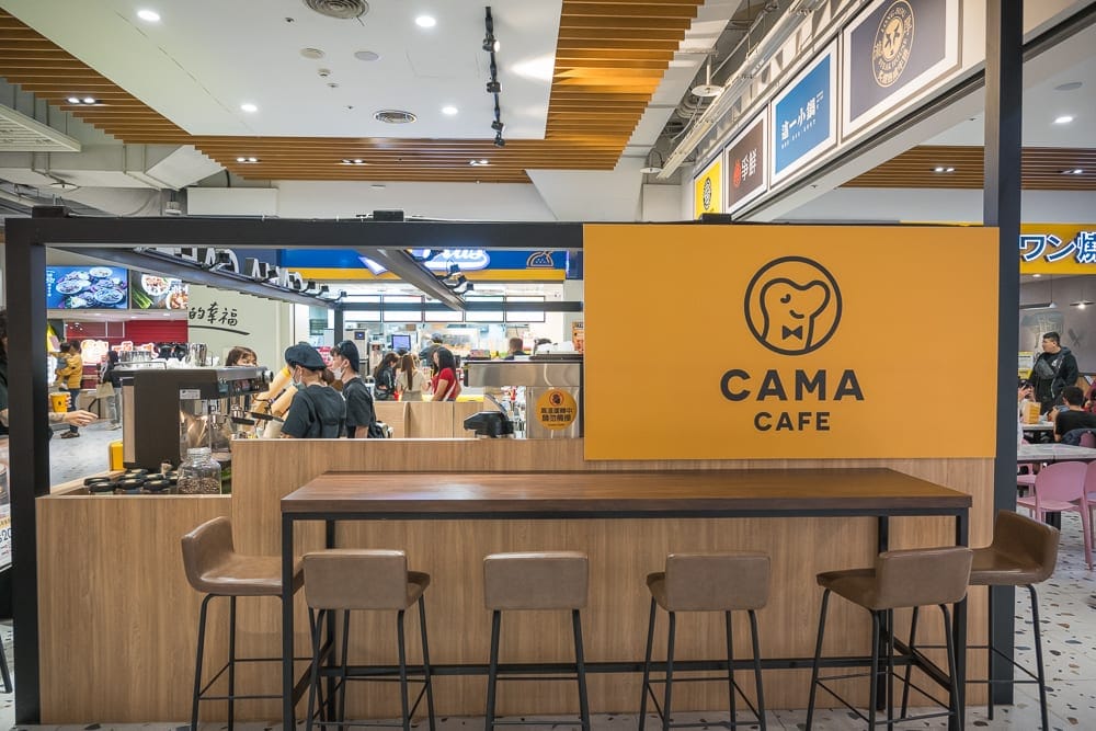 台中大魯閣新時代廣場｜CAMA CAFE 吧台區域，黃色牆面，木質吧台和凳子，現代簡約風格。背景可見其他餐飲店、顧客和員工