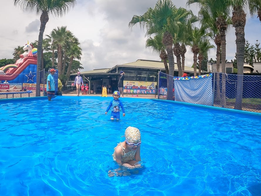 桃園小人國水樂園｜泳池、兒童、泳衣、泳鏡、充氣滑梯、棕櫚樹、建築物、圍欄。