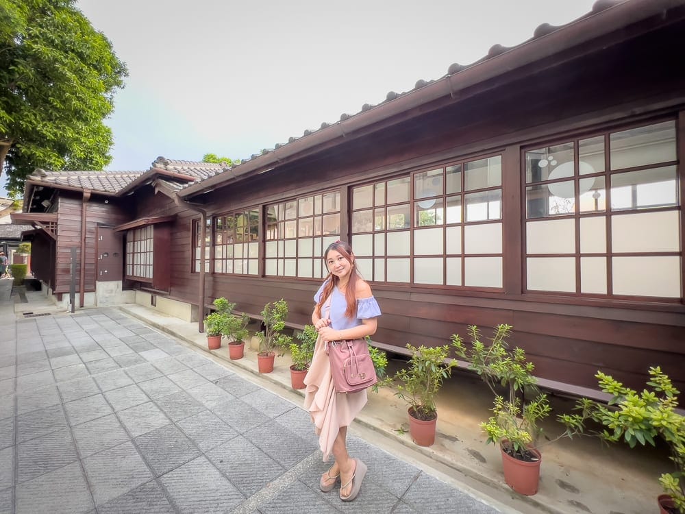桃園大溪老街美食景點｜一座傳統的日式建築、灰色石板路、許多盆栽植物，一名女子穿著休閒的夏季服裝，提著一個粉色包，站在建築物