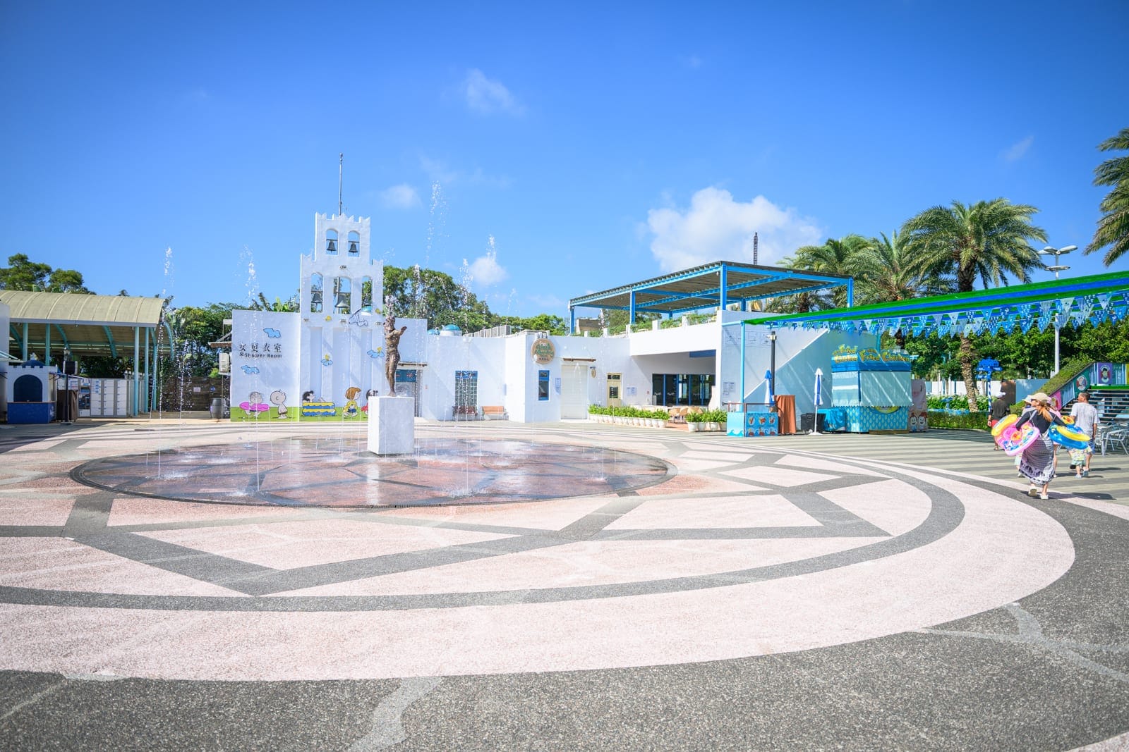 新竹六福水樂園｜廣場、噴泉、建築物、棕櫚樹、遊客。廣場地面有特殊設計，噴泉水流四射，建築物外觀現代且色彩鮮豔，遊客在