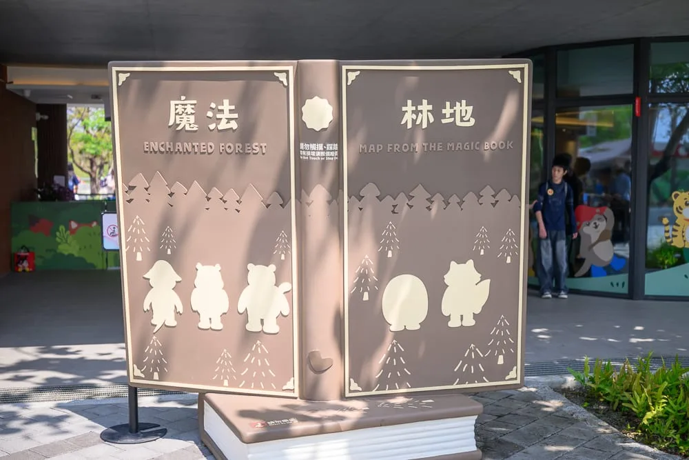 高雄野森動物學校完整攻略|狐狸谷×老鷹小學堂親子玩法解析 11 高雄野森動物學校|大型書本造型看板,寫著魔法林地,樹木圖案,動物剪影,廣場,建築物,人物,戶外。