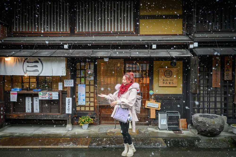 日本岐阜高山三町老街完整攻略|江戶建築群與飛驒牛小吃 13 日本高山-高山老街散冊|女子、街道、雪、木造建築、招牌,飄雪的日式老街,身穿淺色冬衣的女子站在傳統木造店鋪前,雙手微張迎著落