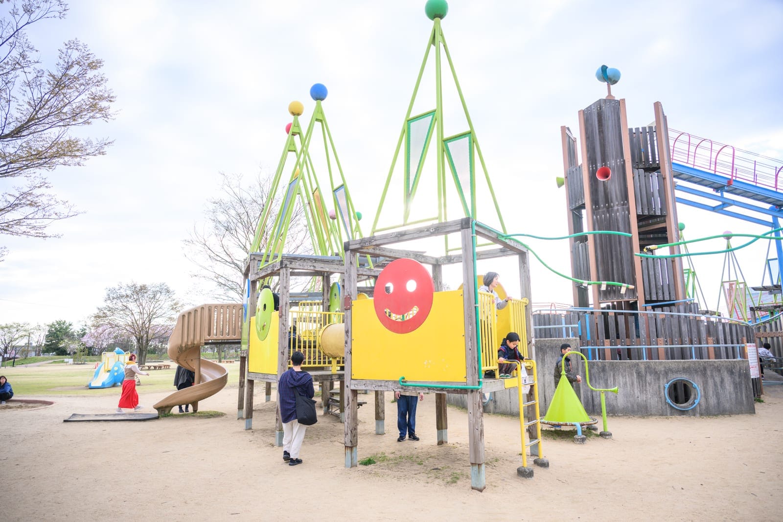 日本富山-高岡童話之森公園完整攻略|哆啦A夢雕塑與親子設施玩法解析 6 日本富山-高岡童話之森公園|戶外公園,大型遊具,溜滑梯,木造塔樓,綠色尖頂支架,沙地,行人,陰天,白天,休閒空間。