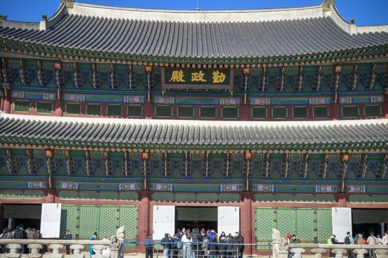 韓國首爾景福宮韓服體驗完整指南|宮殿穿搭租借與親子玩法解析 5 韓國首爾景福宮韓服體驗|傳統宮殿、匾額、飛檐、瓦片、人群,晴空下的宮廷建築外觀,莊嚴而日常的氛圍。