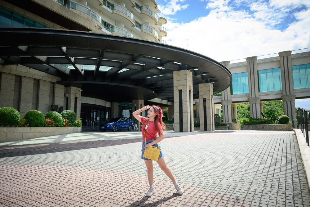 屏東H hotel｜女人，飯店，廣場，藍天，建築物。一名女子站在大型現代飯店前方的寬敞鋪面廣場，右手遮陽、左手提包，建築