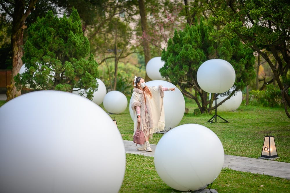 桃園神社｜女子、草地、樹木、白色大圓球、小徑、燈籠。女子站在綠意盎然的戶外草坪與樹林間，四周散落許多大型發光白