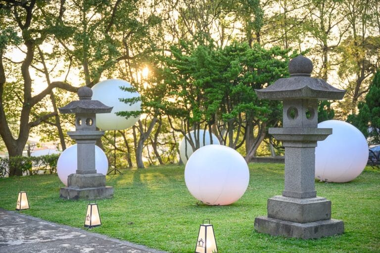 桃園神社_昭和拾參完整攻略|鳥居參道與御守市集玩法解析 18 桃園神社|草地、石燈籠、圓形發光球體、樹林、夕陽,黃昏時刻的戶外庭園散發出柔和光芒,營造出寧靜且神祕的氛圍。