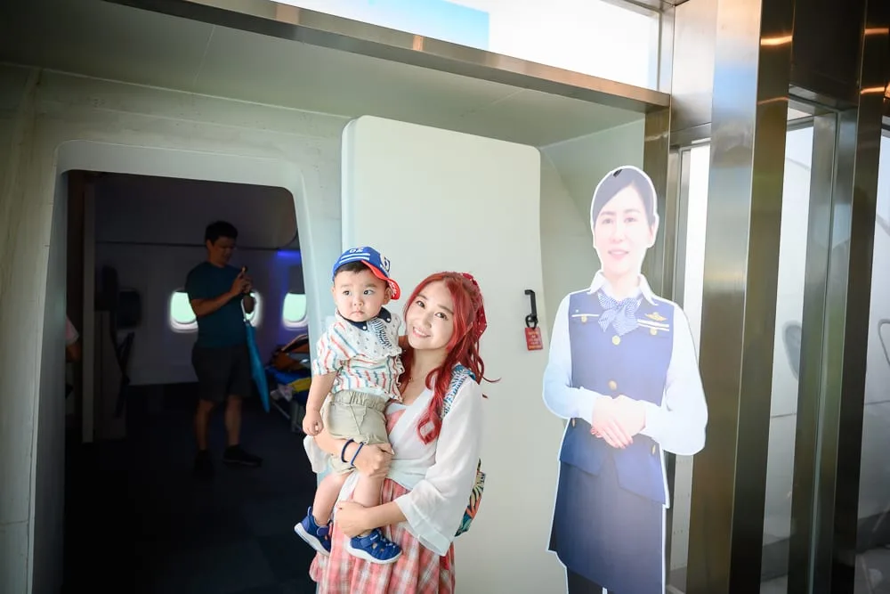 台中中央公園遊客中心完整攻略|機場主題沉浸式展場玩法解析 11 台中中央公園遊客中心|室內,女子,小孩,空服員立牌,飛機艙門。長髮女子抱著戴帽男童站立,旁邊立著穿制服的空服員人形立牌。背