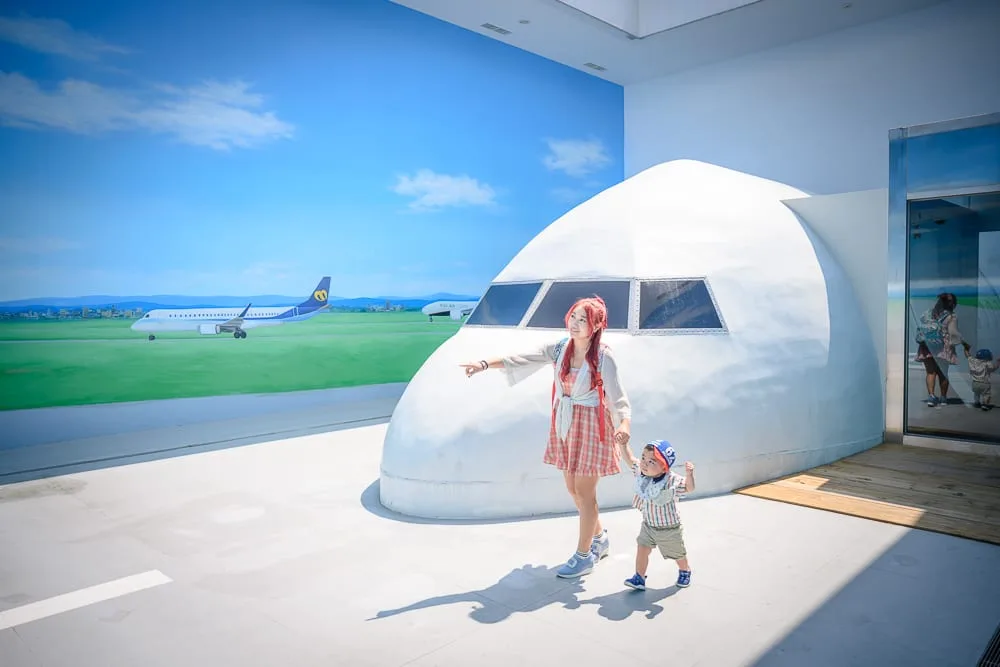 台中中央公園遊客中心完整攻略|機場主題沉浸式展場玩法解析 10 台中中央公園遊客中心|白色建築、飛機造型、機場壁畫、人物、小孩。戶外白色平臺上,一名女子牽著幼童站立,旁側為白色飛機機鼻造