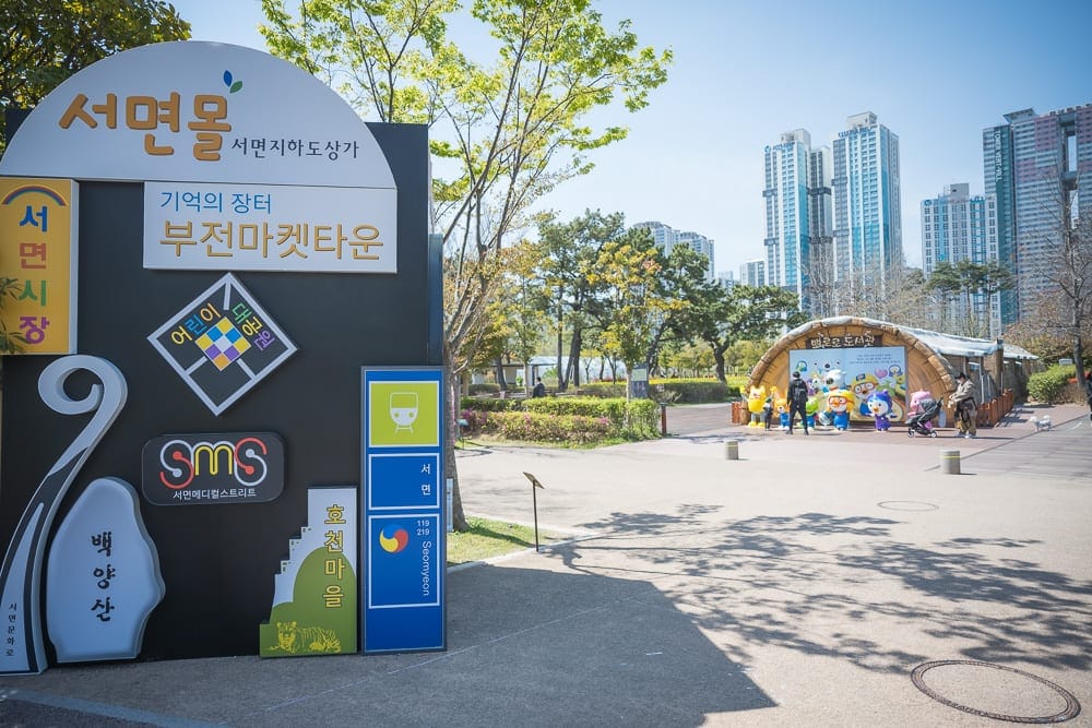 韓國釜山釜山市民公園｜戶外廣場、大型告示牌、綠樹、現代高樓、卡通公仔、晴朗藍天。景觀多元且明亮，背景座落多棟高層建築。