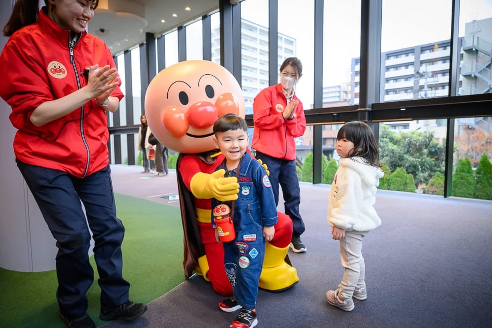 日本仙台-麵包超人博物館｜室內、吉祥物、兒童、工作人員、落地窗，室內明亮空間中，麵包超人吉祥物正與小男孩開心互動，一旁站著小女