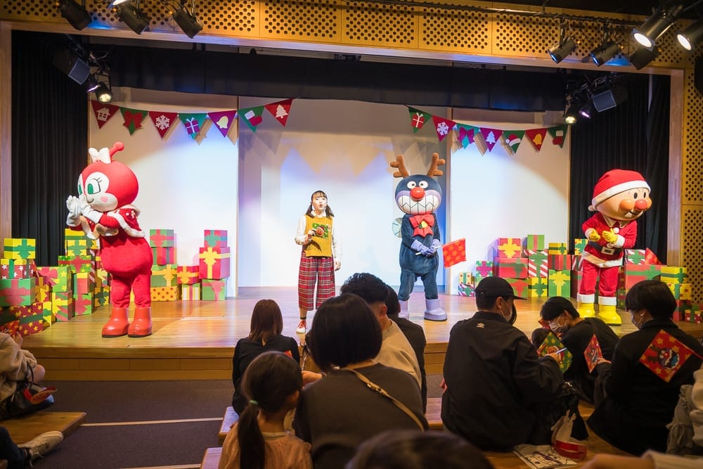 日本仙台-麵包超人博物館｜室內，舞臺，卡通人偶，演員，聖誕裝飾，禮物盒，觀眾。裝飾著彩旗的木質舞臺上，粉紅、藍色與紅色的卡通人