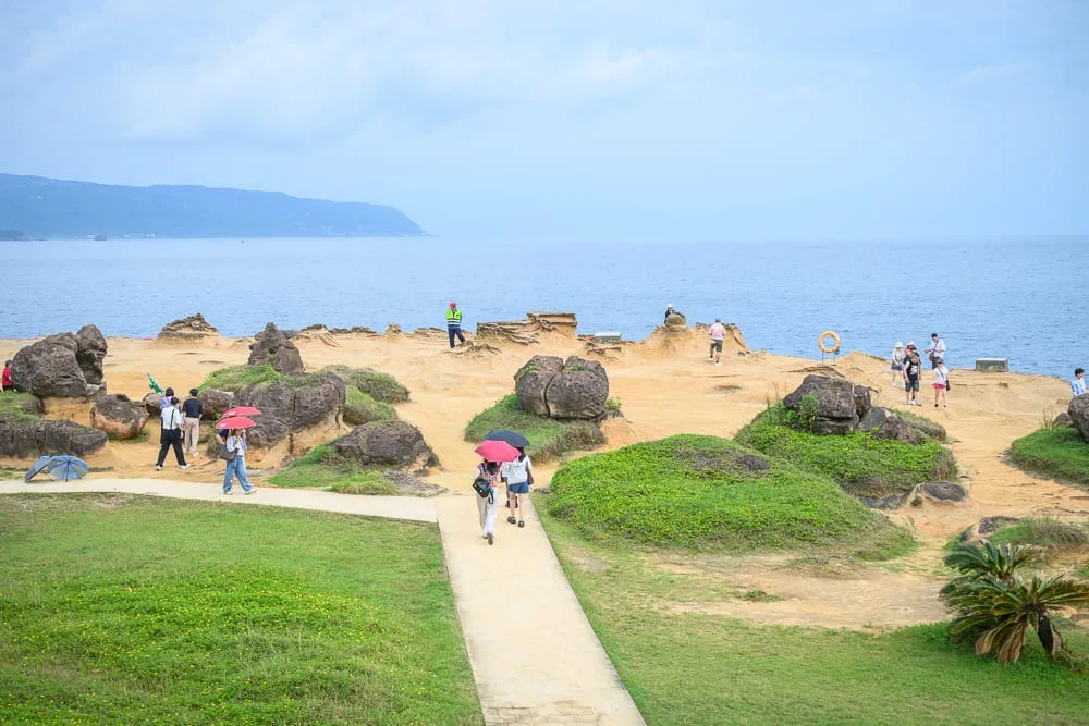 新北野柳地質公園完整攻略|女王頭奇岩怪石玩法解析 10 新北野柳地質公園|海邊,岩石,草地,步道,遊客。蔚藍海岸旁的地質公園內,奇岩怪石羅列,綠意植被點綴。遊客撐傘漫步於步道