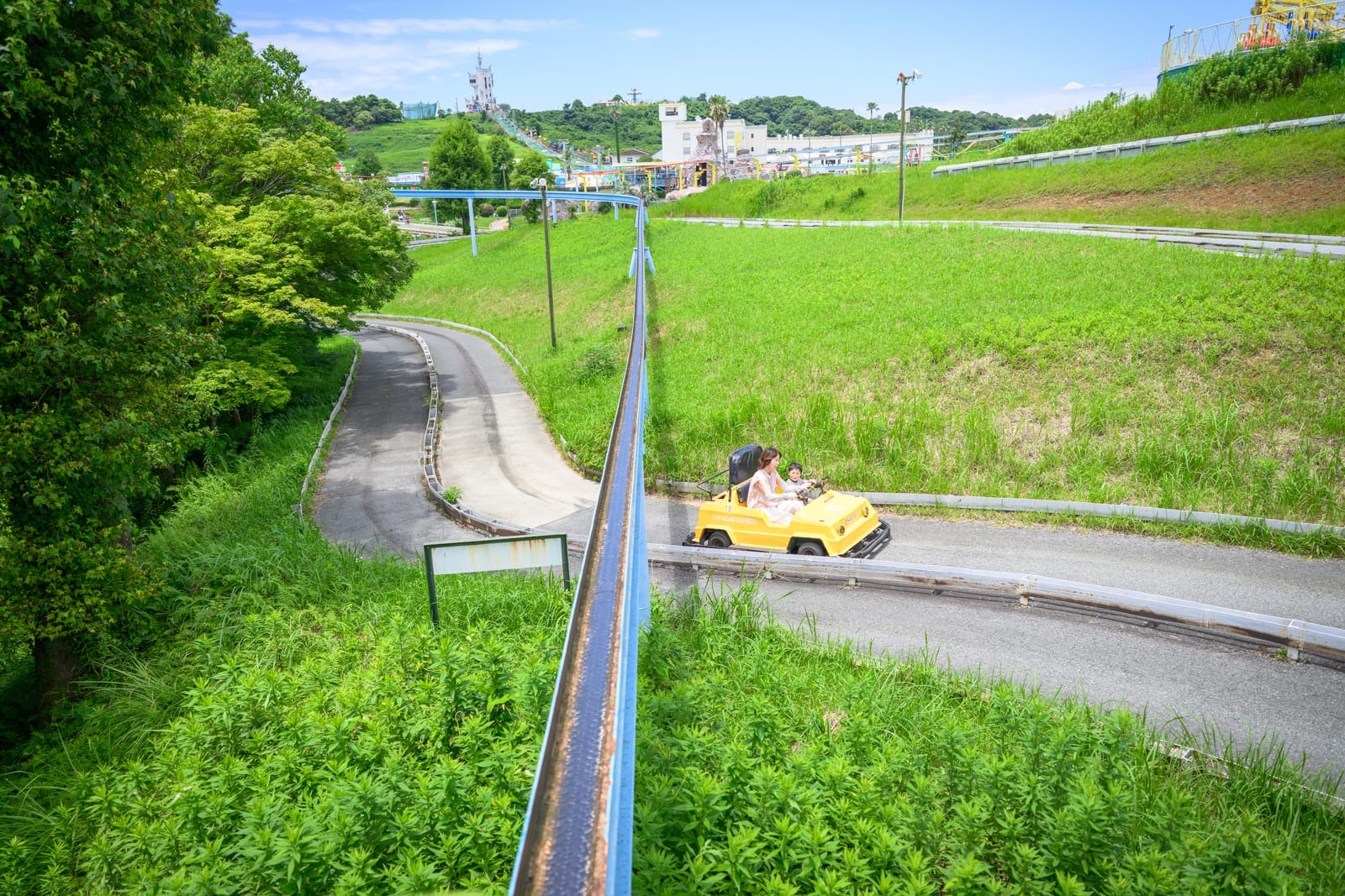 日本熊本-格林主題樂園｜道路、草地、樹、黃色腳踏車、鐵欄杆、建築物、藍天。