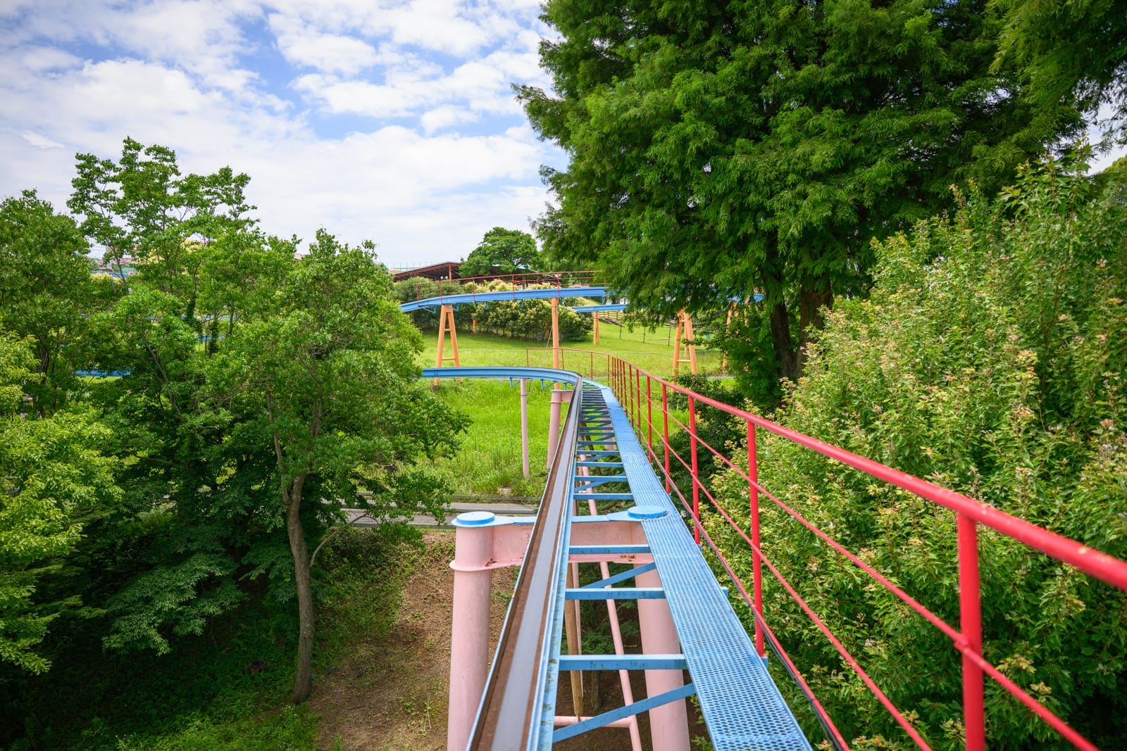 日本熊本-格林主題樂園｜遊樂園軌道、欄杆、樹、草坪、藍天白雲。