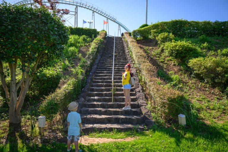 日本熊本-格林主題樂園｜石階、綠草、樹、灌木叢、遊樂園設施、旗幟、藍天，兒童、成人。