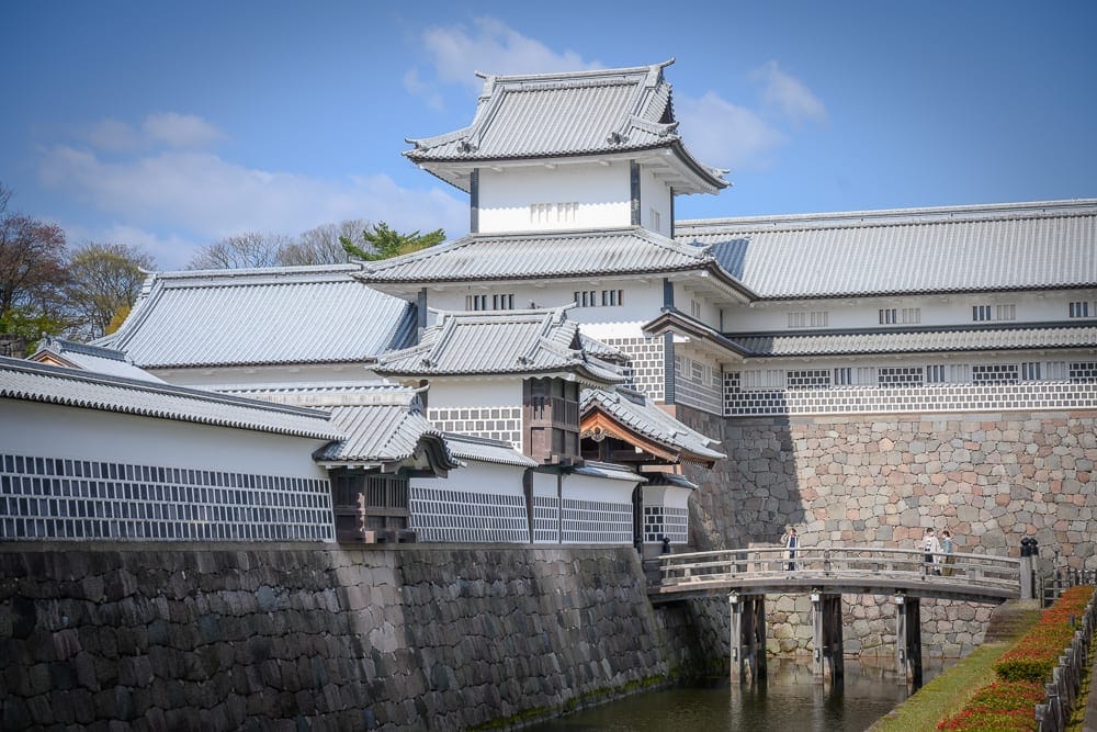 日本金澤-金澤城公園｜藍天下的日式城堡，灰色瓦片屋頂，白牆石垣，木造橋樑，護城河水，幾位遊客佇立橋面。