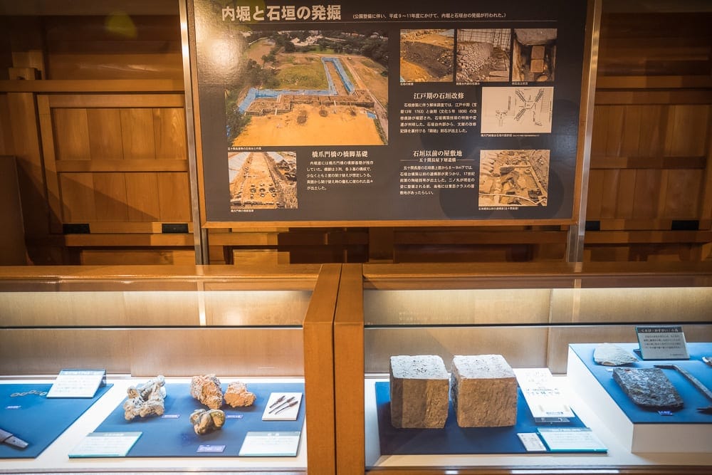 日本金澤-金澤城公園｜室內、展覽、展示櫃、石塊、文物、解說看板、木質牆面。下方玻璃展櫃陳列著石塊、金屬器具與文字說明卡，上