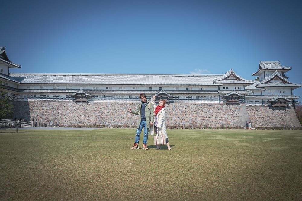 日本金澤-金澤城公園｜城堡、草地、男女、石牆、藍天，一男一女站在綠色草坪中央，背景為白色日式古城與灰色石牆，上方為無雲晴空