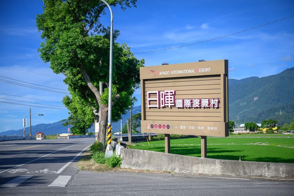 台東日暉渡假村｜藍天白雲下，寬敞的柏油路旁豎立著大型「日暉國際度假村」棕色招牌，一側為綠意盎然的稻田，遠方為層疊的青