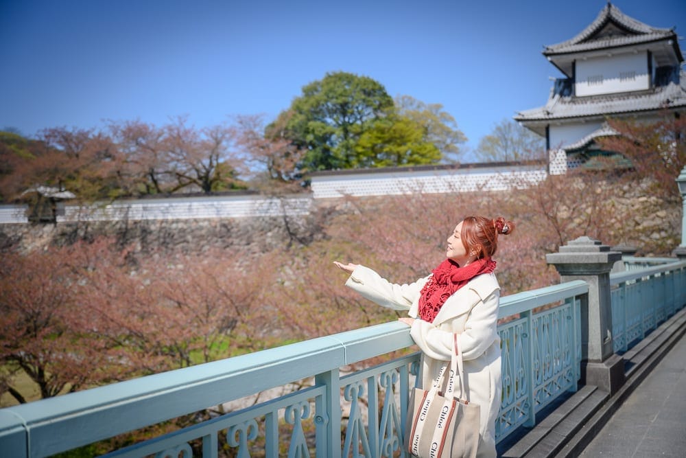 日本金澤-金澤城公園｜女子、橋、城堡、櫻花、藍天。紅髮女子身穿白色大衣與紅圍巾，站在淺藍色欄杆橋樑旁，單手掌心向上平舉。背