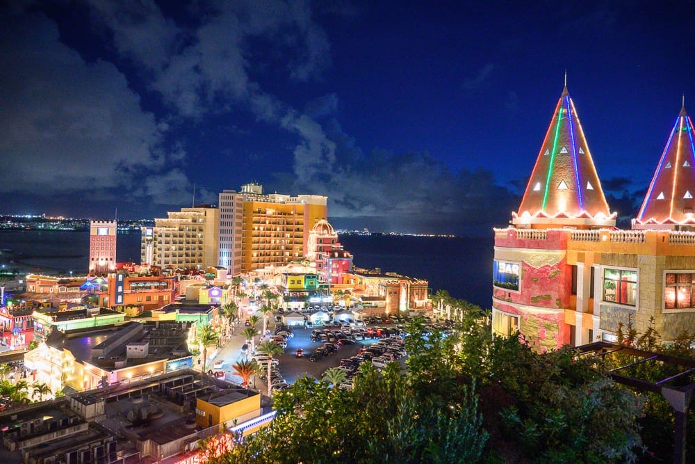 沖繩北谷 LEQU Okinawa 完整攻略|戶外泳池夜景與親子玩法解析 10 沖繩北谷LEQU Okinawa|夜空、海邊、城堡、飯店、燈光、停車場、樹木,夜間海岸邊色彩繽紛的度假村,尖頂建築點綴霓虹燈,遠方海平