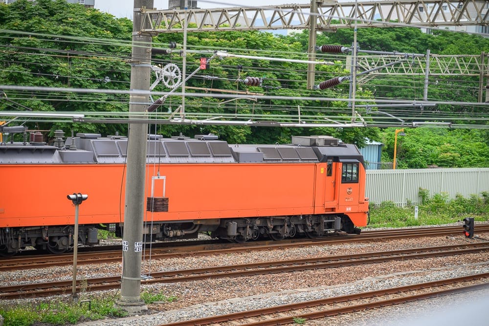 苗栗火車頭園區完整攻略|退役火車頭展示與親子玩法解析 1 苗栗火車頭園區|鐵軌、火車、電線桿、樹木、架空電纜,橘色火車行駛在鐵道上,上方交錯著複雜的電線與金屬支架,背景為綠意