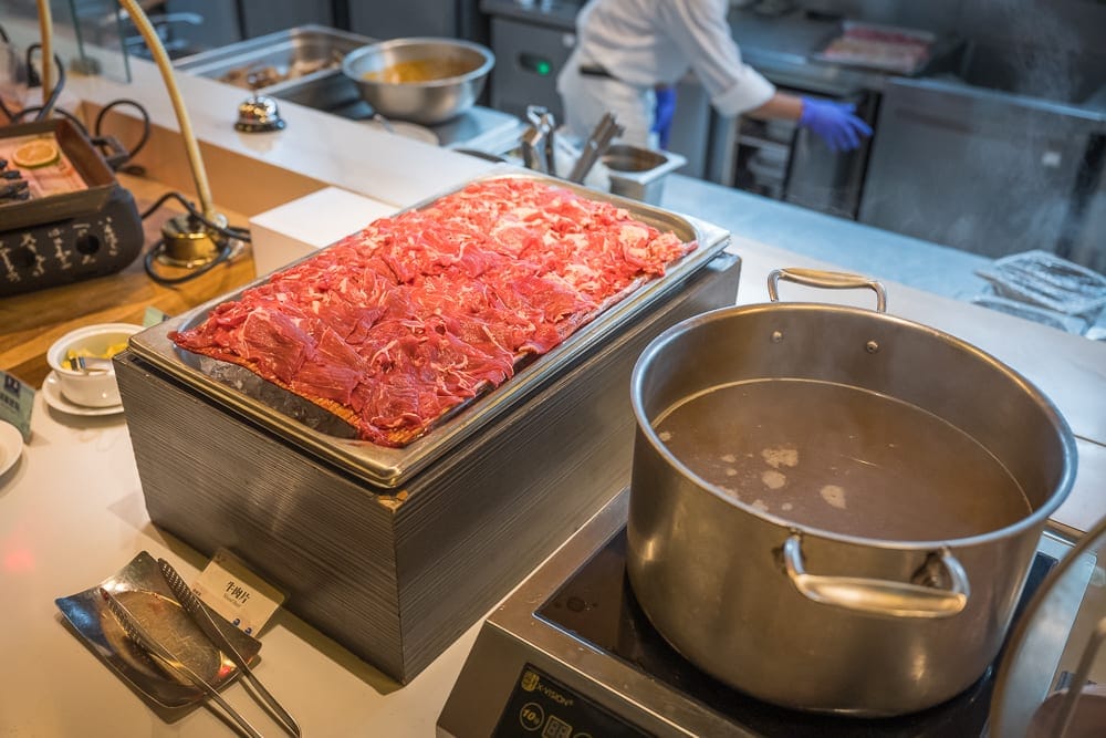 嘉義長榮文苑酒店｜室內、料理、牛肉片、湯鍋、廚房，自助吧檯擺放著切好的牛肉片和一大鍋高湯，背景有廚師在工作，氣氛忙碌。