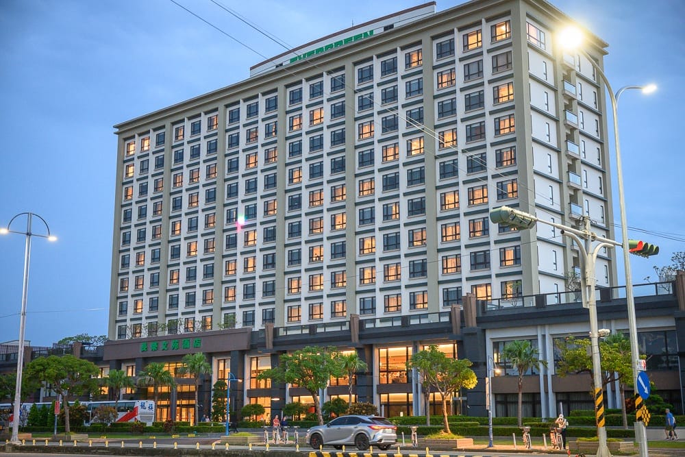 嘉義長榮文苑酒店｜黃昏時刻的高樓建築，飯店，街道，車輛，路燈，天空微藍，窗戶透出溫暖黃光，氛圍平靜。