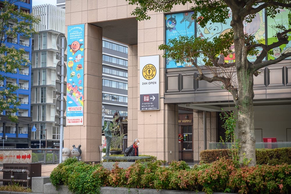 日本福岡麵包超人兒童博物館 in 購物商場完整攻略|親子必玩設施與購票全指南 11 日本福岡-福岡麵包超人博物館|街道,建築物,人,樹木,招牌,卡通海報,行人路,都市景觀,日間,明亮。