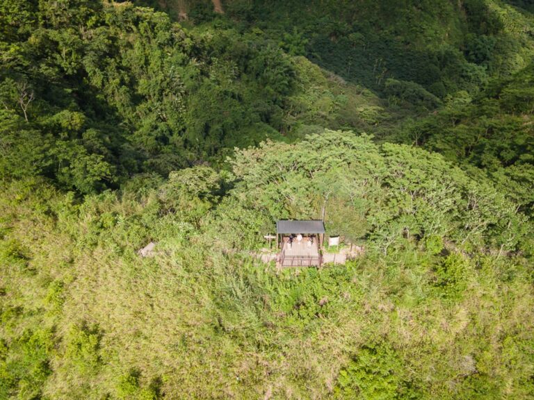 南投九九峰健行步道完整指南|林間土石步道與觀景涼亭玩法解析 11 南投九九峰步道|空拍視角下,群山、茂密樹林與芒草覆蓋山頭,山稜線旁設有一座黑色屋頂的觀景涼亭,數名遊客站立其木棧平台