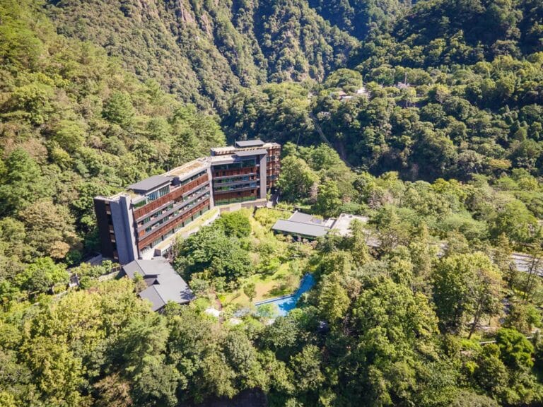 台中統一渡假村 谷關溫泉養生會館完整攻略|親子設施與溫泉泡湯全解析 11 台中谷關統一渡假村|山脈、森林、建築、飯店、游泳池,綠意環繞的山谷中坐落一棟多層樓現代建築,深色外牆搭配大片窗戶,前方空