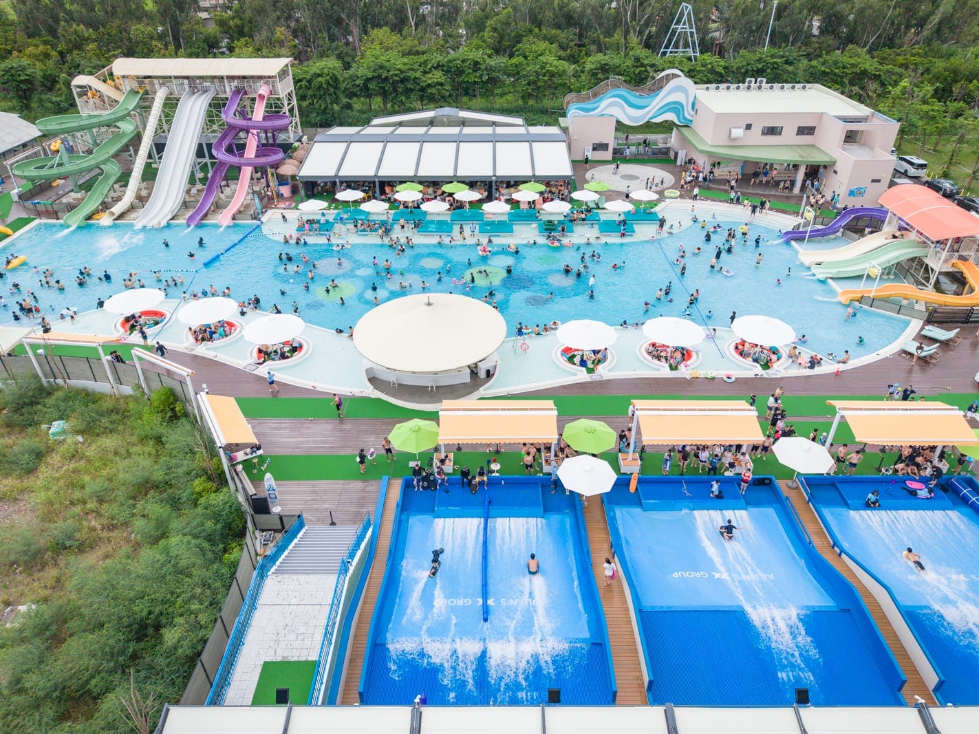 嘉義芯浪國際滑板衝浪公園｜戶外水上樂園，大型游泳池，彩色滑水道，人工造浪池，遮陽傘，樹木，眾多遊客戲水，俯瞰視角