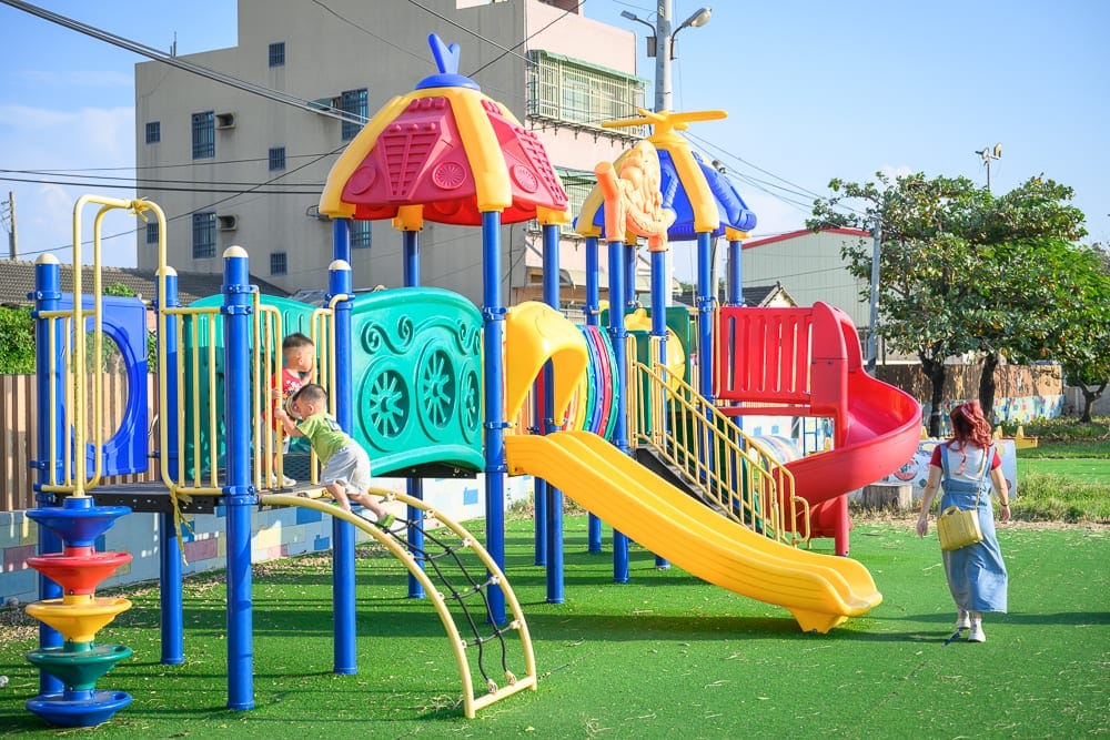 雲林麥寮國小積木公園｜戶外遊樂場，彩色溜滑梯，攀爬架，綠色人工草皮，兩名兒童正在攀爬，一名成人背對行走，背景有建築物與樹木