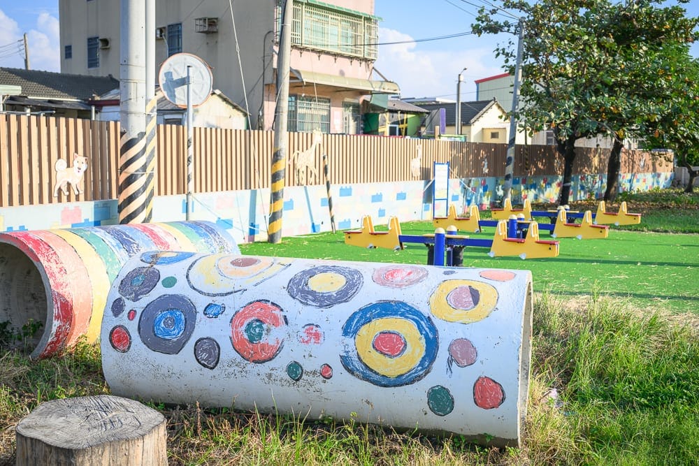雲林麥寮國小積木公園｜戶外公園、草地、彩色水管、翹翹板、木柵欄、樹木、電線桿、民宅。草地上放置著繪有圓形圖案的水管與多組黃