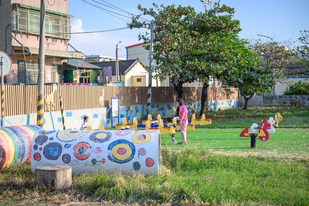 雲林麥寮國小積木公園｜綠地公園、遊樂場、彩色水泥管、搖馬、大人小孩、透天厝、樹木、藍天。成人與幼童在草皮上的遊樂設施旁活動